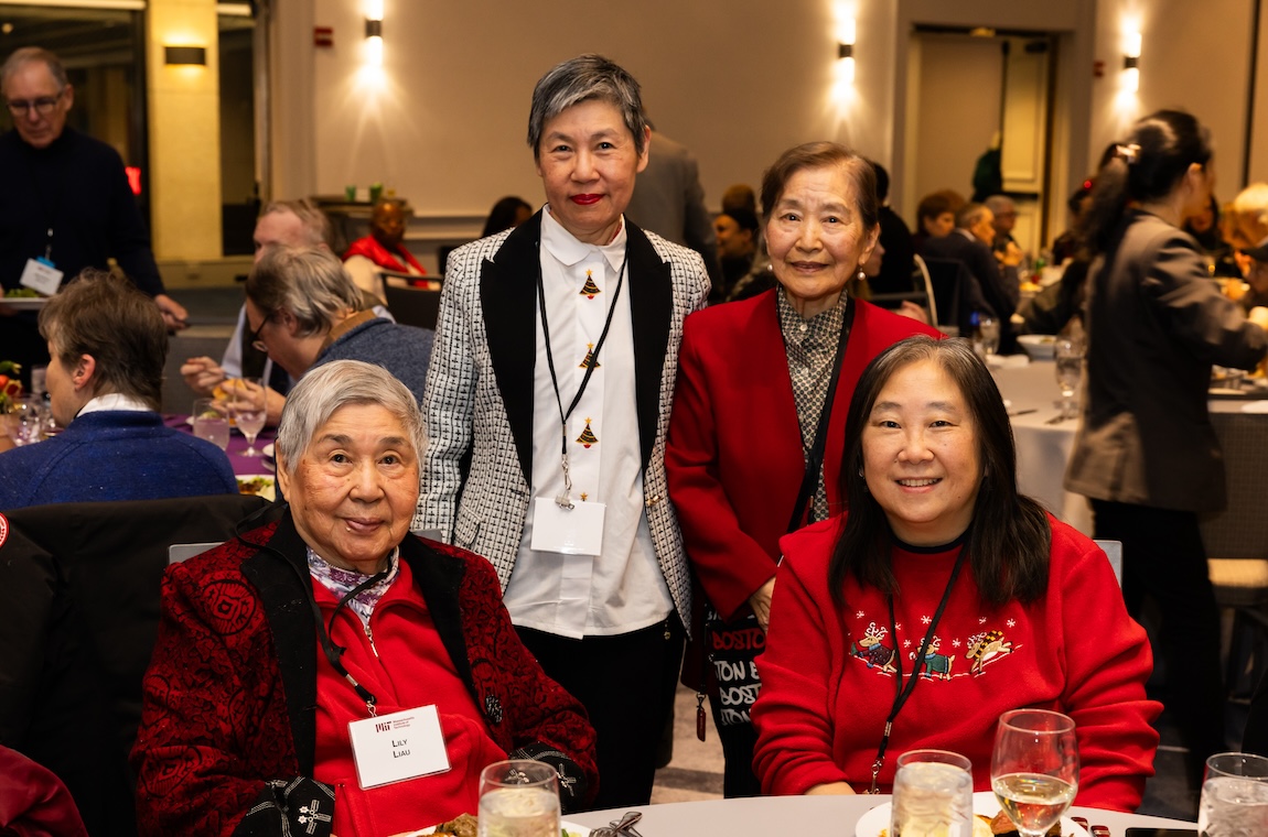 Two members sit at a table while two others stand behind them, all wearing festive holiday outfits and smiling.