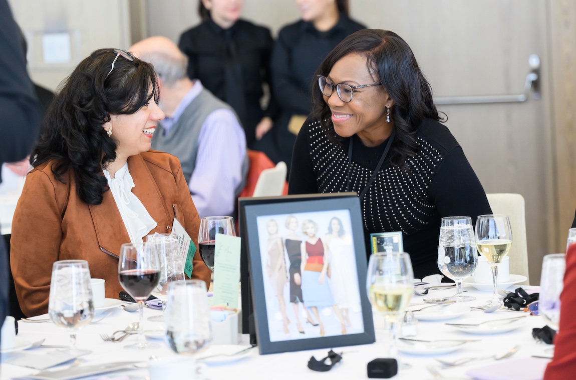 Claudia Forero-Sloan and Maxine Samuels sit at a table and smile. The table is covered by a white tablecloth, plates and wine and water glasses.